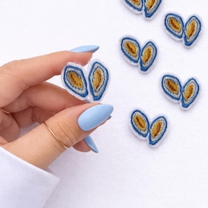May include: Embroidered patches in a heart-like shape, featuring a blue border and a yellow-brown center. Several patches are scattered on a white surface. One patch is held by a hand with blue nail polish.