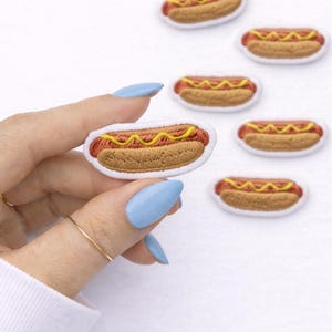 May include: Embroidered hot dog patches with a brown bun, red sausage, and yellow mustard. The patches have a white border. Several patches are scattered on a white background.