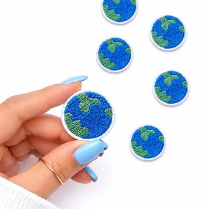 May include: Embroidered patches featuring a blue and green Earth design. The patches have a white border and are round. The image shows several patches, with one held by a hand with a gold ring and blue nail polish.