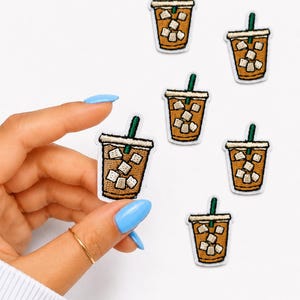 May include: Embroidered patches featuring iced coffee cups with ice cubes and green straws. The patches have a brown cup with a white rim and a tan border. One patch is held by a hand with a gold ring and blue nail polish.
