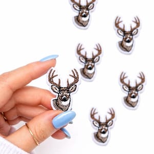 May include: Embroidered deer patches with brown antlers and a detailed face. The patches are white-backed and have a stitched design. One patch is held by a hand with blue nails, and several others are scattered on a white surface.