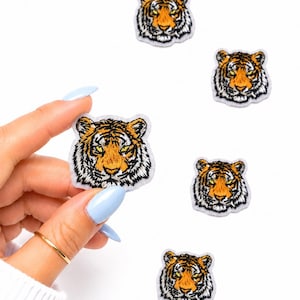 May include: Embroidered tiger head patches with orange, black, and white details. One patch is held by a hand with a gold ring and light blue nails. The patches are on a white background.