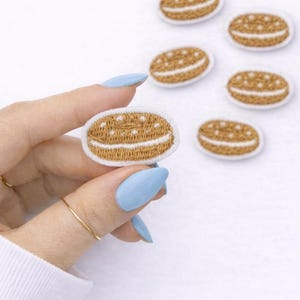 May include: Embroidered patches in the shape of a cookie. The patches are brown with white details and a white border. One patch is held in a hand with blue fingernails, and several others are scattered in the background.