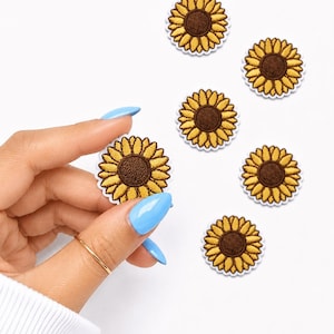 May include: Embroidered sunflower patches with yellow petals and a brown center. One patch is held by a hand with blue fingernails, and several other patches are scattered on a white surface. The patches have a white border.