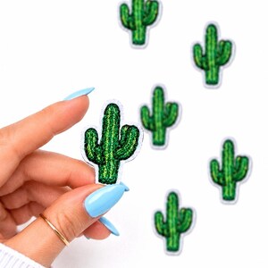 May include: Green cactus patches with white borders. The patches are embroidered with a textured appearance. Several patches are scattered on a white background, with one patch held by a hand with blue fingernails.