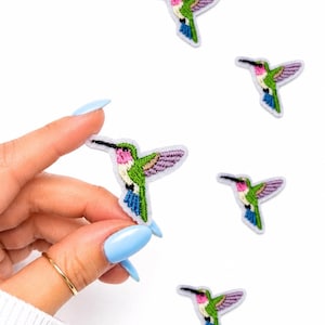 May include: Embroidered hummingbird patches in green, pink, purple, and blue. The patches feature detailed stitching and are set against a white background. One patch is held by a hand with a gold ring and blue nails.