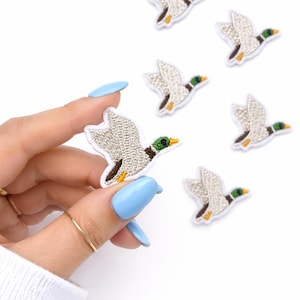 May include: Embroidered duck patches in flight. The patches feature a detailed design with gray wings, green heads, and yellow beaks. The patches are white-bordered and are ideal for adding to clothing or accessories.