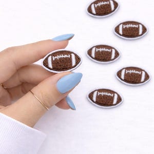 May include: Embroidered football patches in brown and white. The patches are oval-shaped with white stitching and a brown center, resembling a football. Several patches are scattered on a white surface.