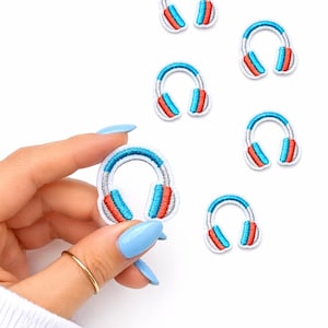 May include: Embroidered patches featuring a headphone design. The patches have a white background with a blue, red, and silver color scheme. The patches are small and suitable for clothing or accessories.