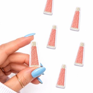 May include: Embroidered patches in the shape of a tube of lip balm. The patches are pink with a white border and a gold-colored cap. One patch is held by a hand with blue fingernails. The patches are scattered on a white background.