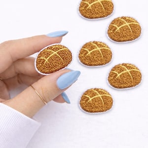 May include: A collection of embroidered patches shaped like a bread roll. The patches are brown with a yellow crosshatch design and a white border. One patch is held by a hand with blue nails.