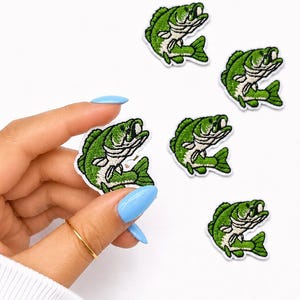 May include: Embroidered patches featuring a green and white bass fish design. The patches are detailed with a white outline and are set against a white background. The image shows several patches, with one held by a hand.