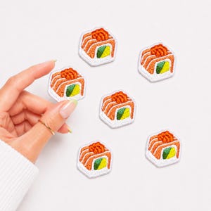 May include: Five embroidered patches shaped like sushi rolls, each featuring orange salmon, a green cucumber center, and white rice, with a textured finish. The patches are arranged in a cluster, with one patch held up to show the scale and detail.