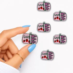 May include: Six small embroidered patches shaped like rectangular tins of sardines. Each patch features a light pink background, three purple fish, two red strawberries, and the text SAR in black thread, all outlined in a white border.