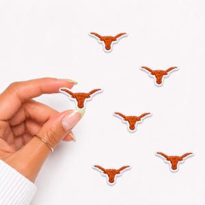May include: Six small embroidered patches featuring a burnt orange longhorn steer head silhouette with a white border. The patches are arranged in a scattered pattern, with one patch being held between fingers to show its small scale and texture.