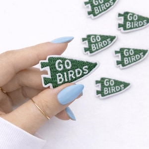 May include: Green and white embroidered patches in an arrowhead shape with the text "GO BIRDS". Several patches are scattered on a white surface. One patch is held by a hand with light blue nail polish.