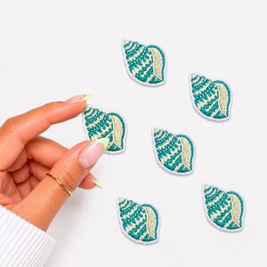 May include: Five embroidered seashell patches featuring a teal and white geometric pattern with a gold-toned edge. Each patch is shaped like a spiral conch shell with detailed stitching throughout the design, showcasing a consistent nautical motif.