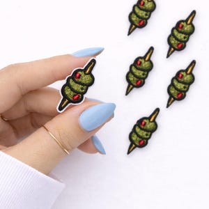 May include: Embroidered olive patches. Each patch features three green olives with red accents, skewered on a gold-colored toothpick. The patches have a black border and are set against a white background.