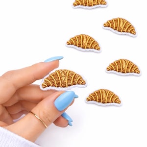 May include: Embroidered patches in the shape of croissants. The patches are brown and yellow with white trim. One patch is held by a hand with blue fingernails, and a gold ring. The patches are on a white background.
