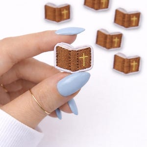 May include: Embroidered patches in the shape of open books with a gold cross on the right page. The patches are brown with white pages and a brown border. Several patches are scattered on a white background.