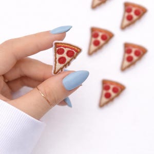 May include: Embroidered pizza slice patches with red pepperoni and a golden crust. The patches are small, with a white border, and are scattered on a white background. A hand holds one patch, with blue nails and a gold ring.