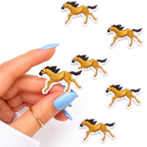 May include: Embroidered patches featuring a running horse design. The patches are tan with black manes and tails, and white borders. One patch is held by a hand with a gold ring and blue nail polish. Several other patches are scattered on a white background.