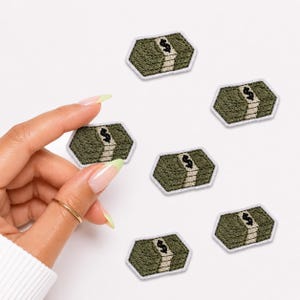 May include: Six embroidered patches shaped like stacks of cash, featuring olive green and beige thread with a black dollar sign symbol on each. The patches have a white border and are arranged in a scattered pattern on a plain white background.