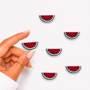 May include: Six small embroidered watermelon slice patches. Each patch features a red curved center with black seed details, a thin white border, and a green outer rind edge. The patches are arranged in a scattered pattern on a plain white background.