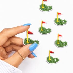 May include: Six embroidered iron-on patches shaped like golf putting greens. Each patch features a green textured surface, a yellow flagstick, and a red flag. The patches are small, circular, and have a white border around the edges.