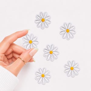 May include: Six embroidered daisy patches featuring white petals and a bright yellow center. Each patch is circular in shape with a stitched border, designed for application onto fabric surfaces as a decorative floral accent for clothing or accessories.
