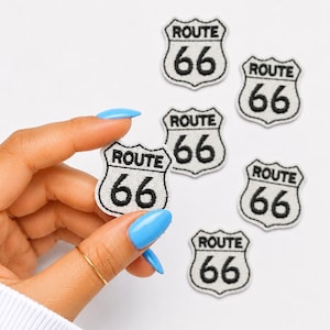 May include: A collection of Route 66 patches. Each patch is white with a black border and features the words "ROUTE 66" above the number "66". One patch is held by a hand with blue nail polish.