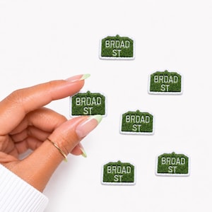 May include: Six small, rectangular, green embroidered patches with white borders and white text that reads BROAD ST. The patches are shaped like street signs with a rounded top edge and are arranged in a scattered pattern on a white background.