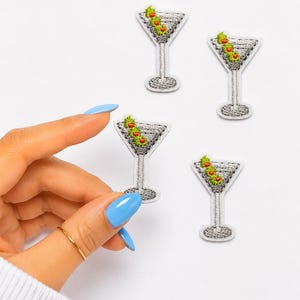 May include: Four embroidered patches shaped like martini glasses. Each patch features a silver-toned glass filled with a grey liquid and three green olives with red centers. The patches have a white border and a textured, stitched appearance.