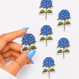 May include: Six embroidered patches featuring blue hydrangea flower heads with green stems and leaves. Each patch has a white border. The floral design is rendered in shades of blue and green thread, creating a textured appearance for the petals.