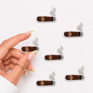 May include: A collection of six embroidered iron-on patches, each depicting a brown cigar with a tan band and a wisp of grey smoke rising from the end. The patches are arranged in a scattered pattern against a plain white background.