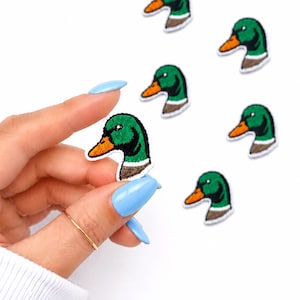 May include: Embroidered duck head patches with green, orange, and brown details. The patches are small and feature a detailed design of a duck's head. Several patches are scattered on a white background.