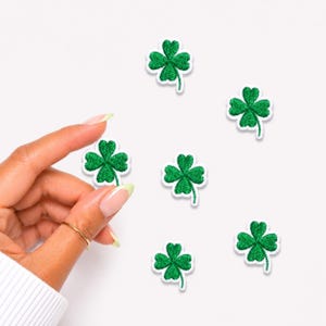 May include: Five small, embroidered four-leaf clover patches in a vibrant green color with white stitched borders. The clover shapes are arranged in a scattered pattern, with one patch being held up to display the detailed texture of the thread.