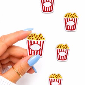 May include: Embroidered popcorn patches. Each patch features a red and white striped popcorn container filled with yellow popcorn. A hand holds one patch, with others scattered on a white background.