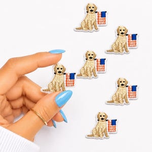 May include: Six embroidered patches featuring a golden-colored dog sitting next to a small American flag. Each patch depicts a tan dog with a black nose and eyes, positioned beside a rectangular flag with red and white stripes and a blue square.