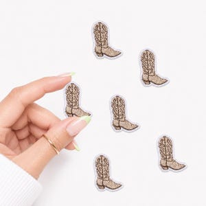 May include: A collection of six small, embroidered cowboy boot patches. Each patch features a tan and brown color scheme with detailed stitching that mimics the texture of leather. The boots are oriented in various directions across the frame.