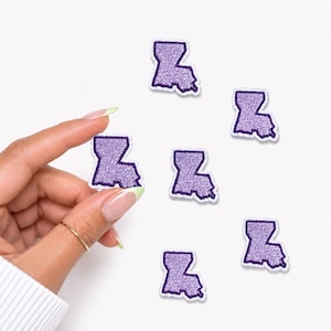May include: Six small, purple embroidered patches in the shape of the state of Louisiana. Each patch features a textured, light purple interior with a dark purple stitched border, creating a distinct outline of the state's geographic shape.