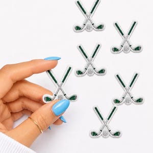 May include: Five embroidered iron-on patches featuring a pair of crossed golf clubs with green shafts and silver heads, centered above a small silver golf ball. The patches are white with a glittery texture, showcasing a detailed sports theme.