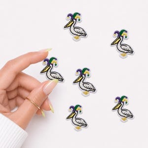 May include: Six small embroidered patches, each featuring a white pelican wearing a jester hat in purple, green, and yellow. The pelicans have yellow beaks and feet, with black outlines defining their shapes. The patches are arranged on a plain surface.