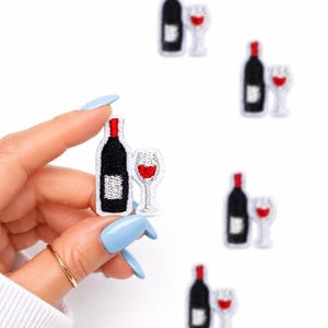May include: Embroidered patches featuring a black wine bottle with a red top and a wine glass with red liquid. The patches are white, black, and red. The image shows a hand holding one patch, with several others scattered around.