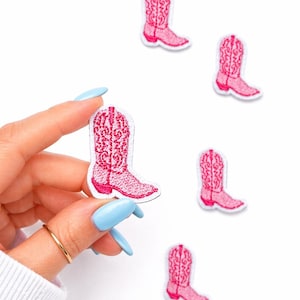 May include: Pink embroidered cowboy boot patches with intricate detailing. The patches are in a vibrant pink color and have a white border. Several patches are scattered on a white background.