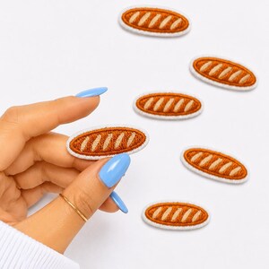 May include: A collection of six oval-shaped embroidered patches, each featuring a brown baguette design with diagonal cream-colored score lines and a white stitched border. The patches are arranged in a scattered pattern on a plain white background.