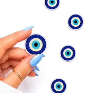 May include: Embroidered patches featuring a blue and white evil eye design. The patches have a circular shape with a dark blue outer ring, a light blue inner ring, a yellow ring, and a black center. The patches are on a white background.
