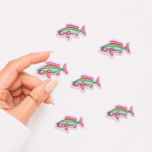 May include: A collection of six small embroidered fish patches. Each patch features a vibrant design with a bright turquoise body, a shimmering gold midsection, and bold pink outlines on the fins, tail, and dorsal area, with a small blue eye.