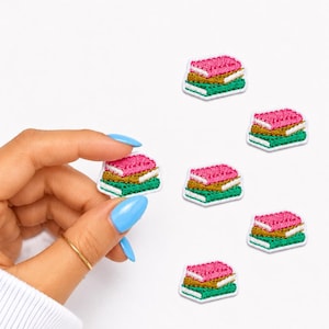 May include: Embroidered patches in the shape of stacked books. Each patch features a pink top, gold middle, and green bottom, all outlined in white. The patches are scattered on a white background.