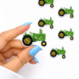 May include: Embroidered green tractor patches with black and yellow wheels. The patches are on a white background. One patch is held by a hand with blue nail polish and a gold ring.